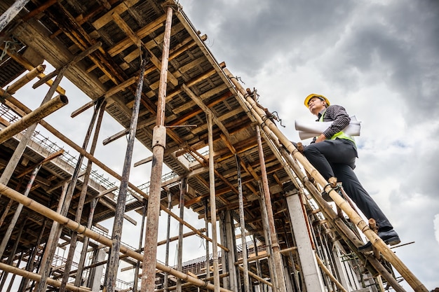 Jeune ingénieur professionnel travailleur dans le casque de protection et le papier des plans sur place travaillant sur l'échelle sur le chantier de construction de la maison