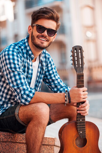 Jeune et insouciant. Souriant jeune homme tenant une guitare et regardant la caméra pendant que l'homme est assis à l'extérieur