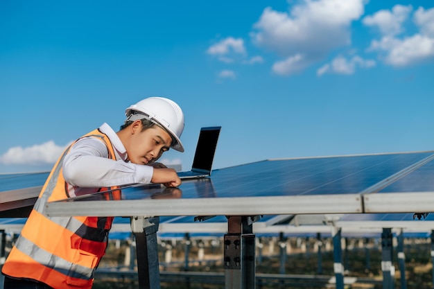 Jeune inspecteur asiatique Ingénieur homme utilise un ordinateur portable travaillant à la ferme solaire Technicien superviseur mâle en casque blanc Vérification du fonctionnement du soleil et du panneau solaire photovoltaïque dans l'espace de copie de la station