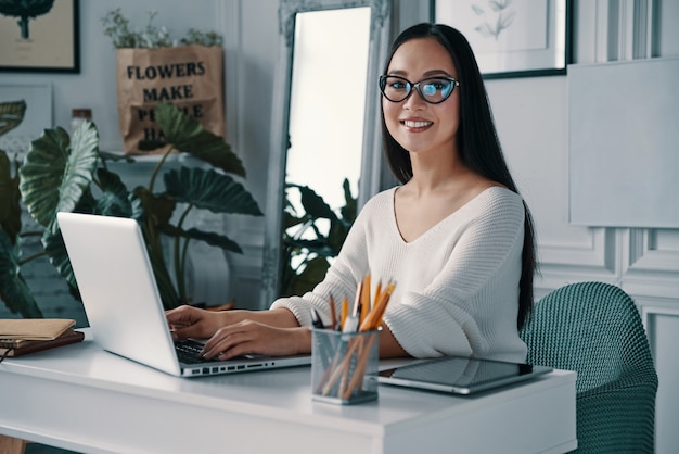 Jeune et intelligent. Belle jeune femme souriante et regardant la caméra alors qu'elle était assise au bureau à domicile