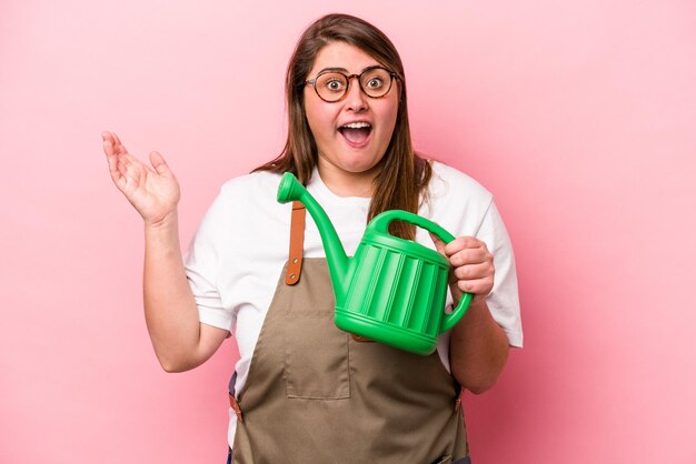 Jeune jardinier caucasien en surpoids femme tenant un arrosoir fond isolé recevant une agréable surprise, excité et levant les mains.