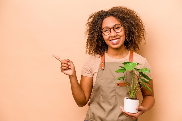 Jeune jardinier femme afro-américaine tenant une plante isolée sur fond beige souriant et pointant de côté montrant quelque chose à l'espace vide
