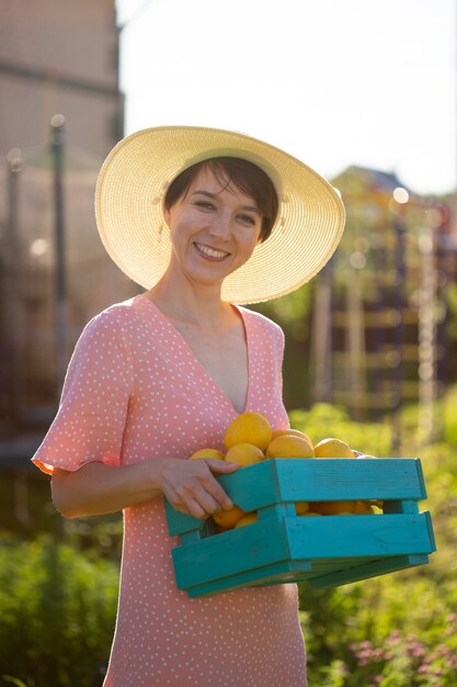 Jeune jardinier de femme positive caucasienne en chapeau et robe rose tient la boîte avec des citrons dans ses mains sur la journée d'été ensoleillée. Concept d'agriculture et de jardinage biologique