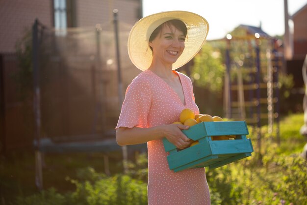 Jeune jardinier de femme positive caucasienne en chapeau et robe rose tient la boîte avec des citrons dans ses mains sur la journée d'été ensoleillée. Concept d'agriculture et de jardinage biologique