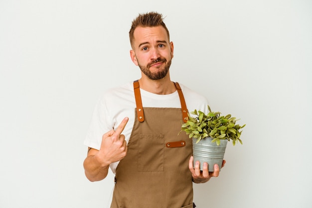 Jeune jardinier tatoué homme caucasien tenant une plante isolée sur fond blanc pointant avec le doigt sur vous comme si vous invitiez à vous rapprocher.