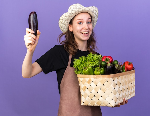 Jeune jardinière impressionnée portant un chapeau de jardinage tenant un panier de légumes élevant des aubergines