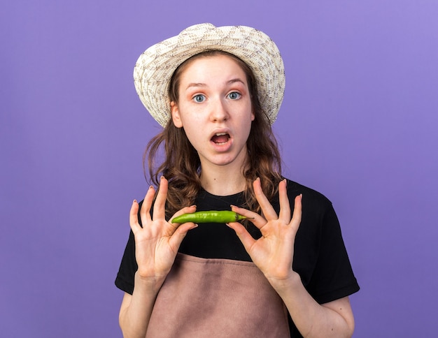 Jeune jardinière surprise portant un chapeau de jardinage tenant du poivre isolé sur un mur bleu