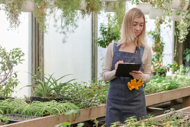 Une jeune jardinière tient un registre des fleurs dans une serre au printemps.