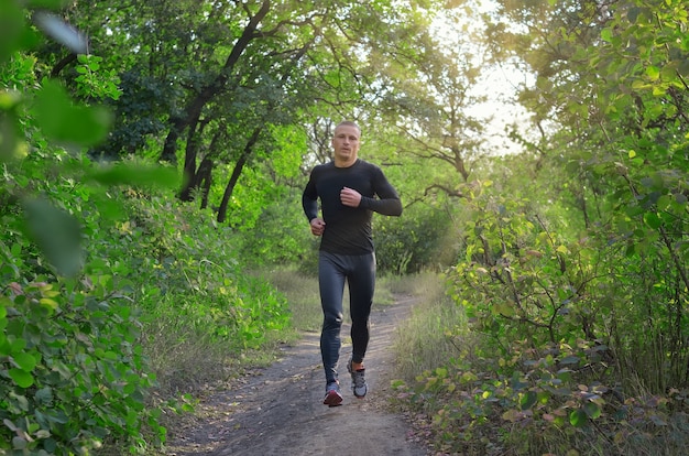 Un jeune jogger fort vêtu de leggings de sport noirs, d'une chemise et de baskets court sur la forêt verte du printemps. La photo montre un mode de vie sain.