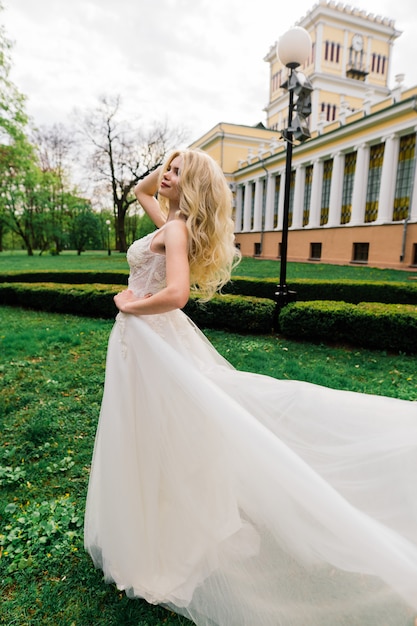 Jeune jolie blonde mariée aux cheveux bouclés marchant dans le parc et souriant