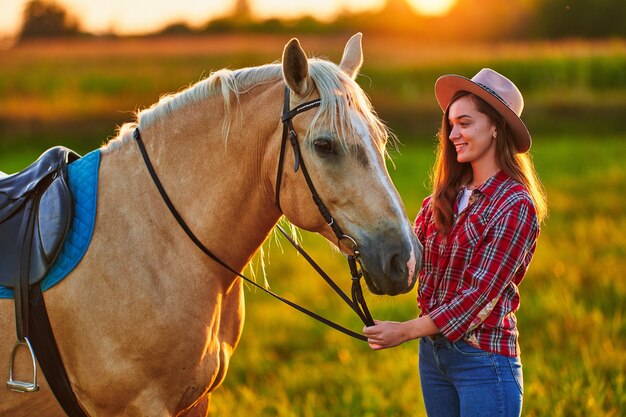 Jeune jolie cow-girl souriante heureuse joyeuse satisfaite avec un beau cheval palomino blond au pré au coucher du soleil