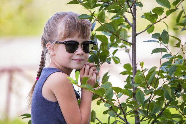 Jeune jolie enfant fille dans des lunettes de soleil à la mode étreignant petit arbre vert en été à l'extérieur.