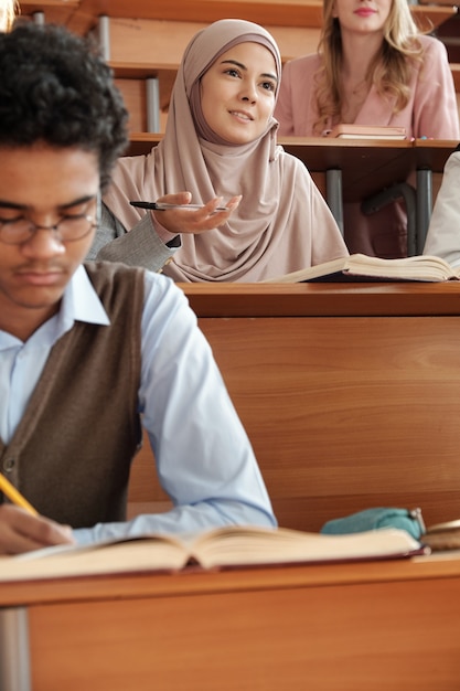 Jeune jolie étudiante en hijab assise à son bureau pendant la conférence et regardant l'enseignant tout en l'écoutant parmi ses camarades de groupe