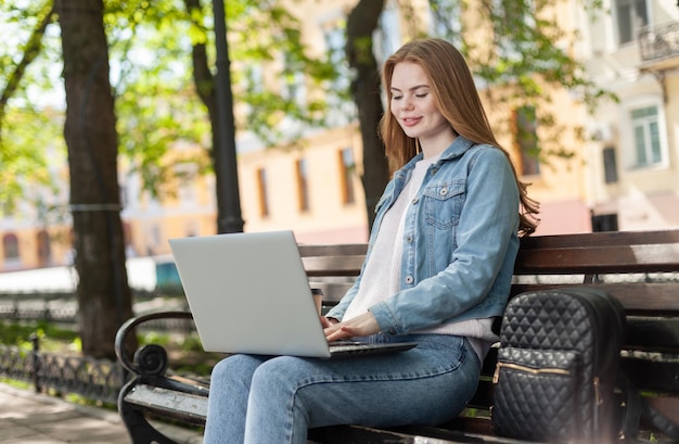 Jeune jolie étudiante ou pigiste utilisant un ordinateur portable assis sur un banc dans la ville