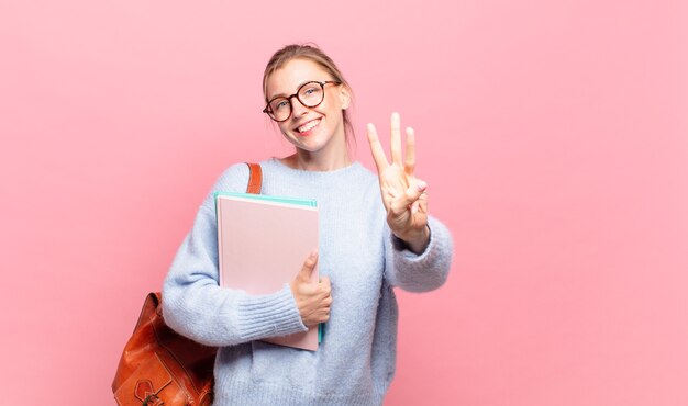 Jeune jolie étudiante souriante et semblant amicale, montrant le numéro trois ou troisième avec la main en avant, comptant à rebours