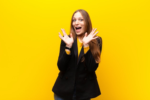 Jeune jolie femme d'affaires air heureux et excité, choqué par une surprise inattendue avec les deux mains ouvertes à côté de face sur le mur orange