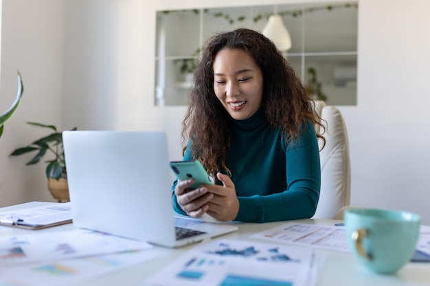 Jeune jolie femme d'affaires asiatique au bureau assise sur une chaise de bureau à l'aide de son smartphone