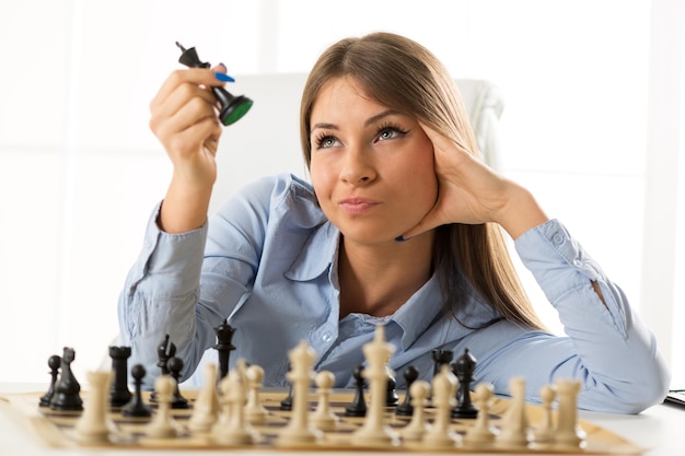 Jeune jolie femme d'affaires assise à la table sur un échiquier reposant sur sa main en regardant la pièce d'échecs du roi noir qui tient dans l'autre main.