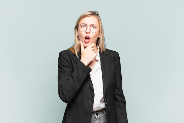 Jeune jolie femme d'affaires avec la bouche et les yeux grands ouverts et la main sur le menton, se sentant désagréablement choquée, disant quoi ou wow