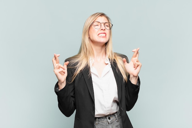 Jeune jolie femme d'affaires croisant les doigts avec anxiété et espérant bonne chance avec un regard inquiet