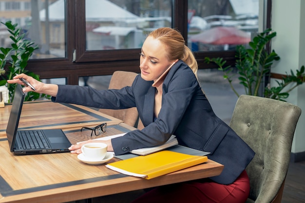 Jeune jolie femme d'affaires parler au smartphone et à la recherche d'ordinateur portable pour travailler au bureau