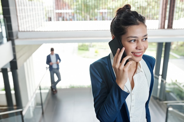 Jeune jolie femme d'affaires positive debout dans un immeuble de bureaux et parlant au téléphone