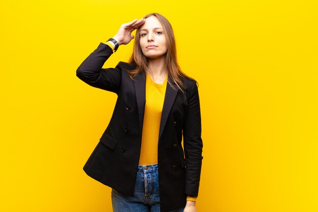 Jeune jolie femme d'affaires saluant l'appareil photo avec un salut militaire dans un acte d'honneur et de patriotisme, montrant du respect sur fond orange