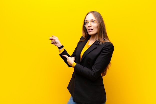 Jeune jolie femme d'affaires se sentant choquée et surprise, montrant du doigt l'espace de copie avec un regard étonné et bouche bée contre le mur orange