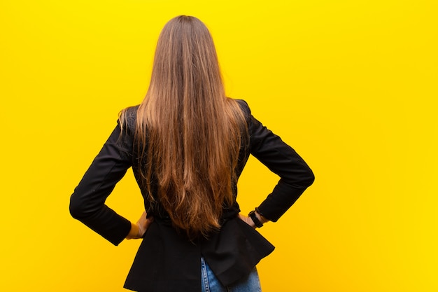 Jeune jolie femme d'affaires se sentant confuse ou pleine ou des doutes et questions, se demandant, avec les mains sur les hanches, vue arrière contre orange