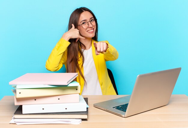 Jeune jolie femme d'affaires souriant joyeusement et pointant vers la caméra tout en faisant un appel, vous geste plus tard, parler au téléphone