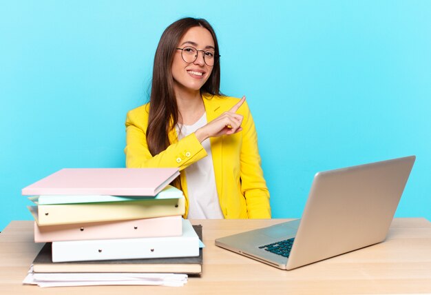 Jeune jolie femme d'affaires souriant joyeusement, se sentant heureux et pointant vers le côté et vers le haut, montrant l'objet dans l'espace de copie