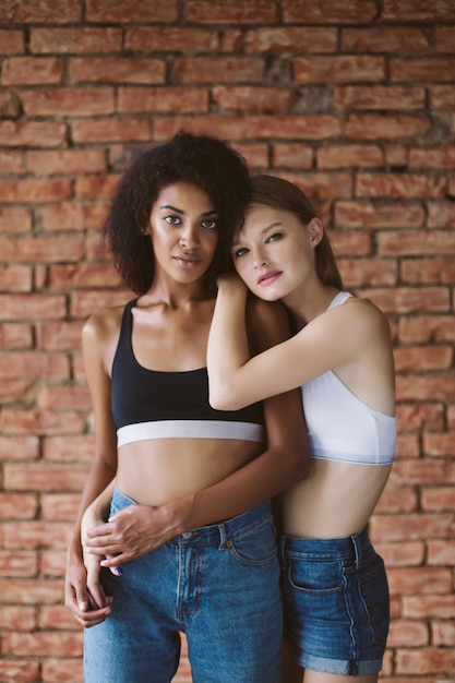 Jeune jolie femme afro-américaine aux cheveux bouclés foncés et belle femme aux cheveux blonds rêveusement avec mur de briques isolé