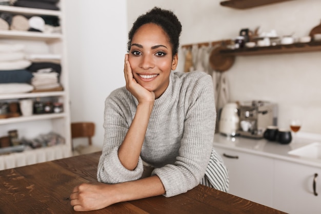 Jeune jolie femme afro-américaine souriante aux cheveux bouclés foncés, s'appuyant sur la table avec bonheur