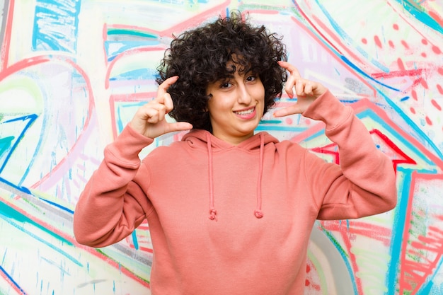 Jeune jolie femme afro encadrant ou décrivant son propre sourire à deux mains, à la recherche positive et heureuse, bien-être contre un mur de graffitis