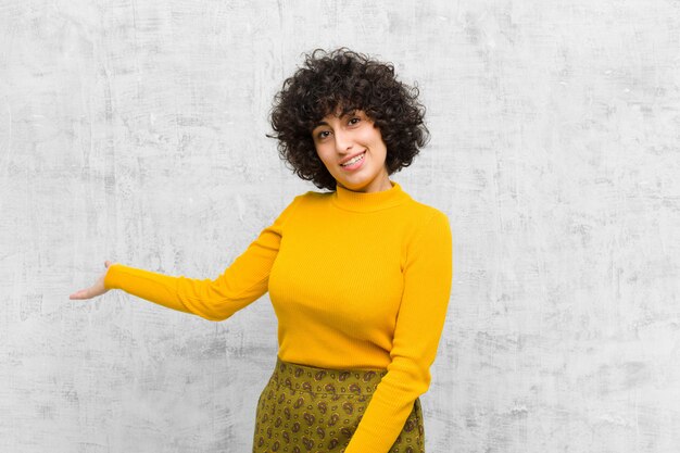 Jeune jolie femme afro se sentant heureuse et joyeuse, souriante et vous accueillant, vous invitant avec un geste amical