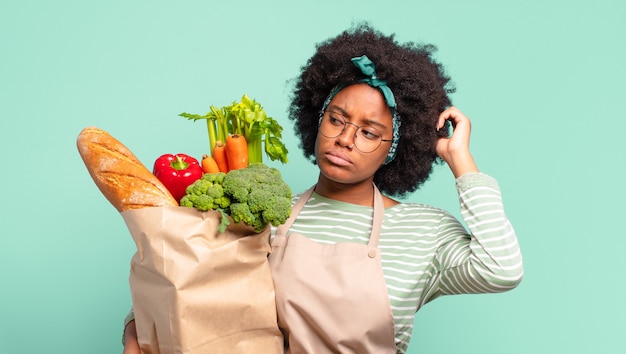 Jeune jolie femme afro souriant joyeusement et rêvassant ou doutant, regardant sur le côté et tenant un sac de légumes