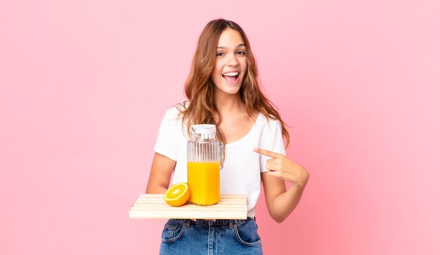 Jeune jolie femme à l'air excitée et surprise pointant sur le côté et tenant un plateau avec un jus d'orange