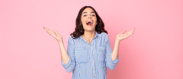 Jeune jolie femme à l'air heureuse et excitée, choquée par une surprise inattendue avec les deux mains ouvertes à côté du visage