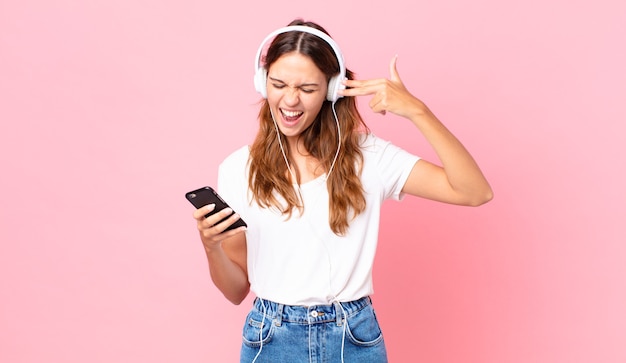 Jeune jolie femme à l'air malheureuse et stressée, geste de suicide faisant un signe d'arme à feu avec des écouteurs et un smartphone