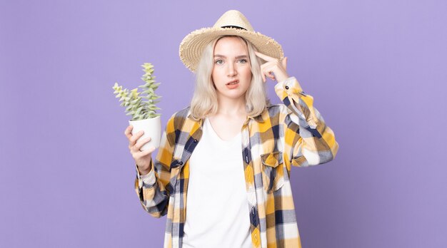 Jeune jolie femme albinos se sentant confuse et perplexe, vous montrant que vous êtes fou et tenant un cactus de plante d'intérieur