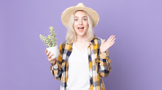 Jeune jolie femme albinos se sentant heureuse et étonnée de quelque chose d'incroyable et tenant un cactus de plante d'intérieur