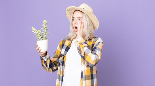 Jeune jolie femme albinos se sentant heureuse, excitée et surprise et tenant un cactus de plante d'intérieur