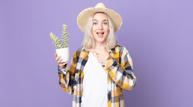 Jeune jolie femme albinos semblant excitée et surprise en pointant sur le côté et tenant un cactus de plante d'intérieur