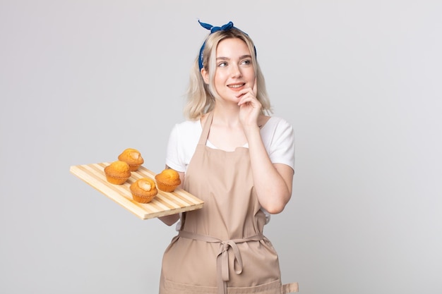 Jeune jolie femme albinos souriant joyeusement et rêvant ou doutant avec un plateau à muffins