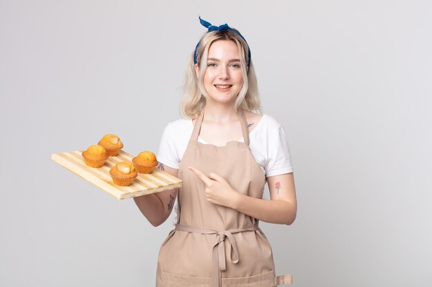 Jeune jolie femme albinos souriant joyeusement se sentant heureuse et pointant vers le côté avec un plateau de muffins