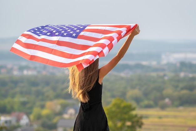 Jeune jolie femme américaine aux cheveux longs tenant en agitant le vent drapeau américain sur ses épaules debout à l'extérieur en profitant d'une chaude journée d'été