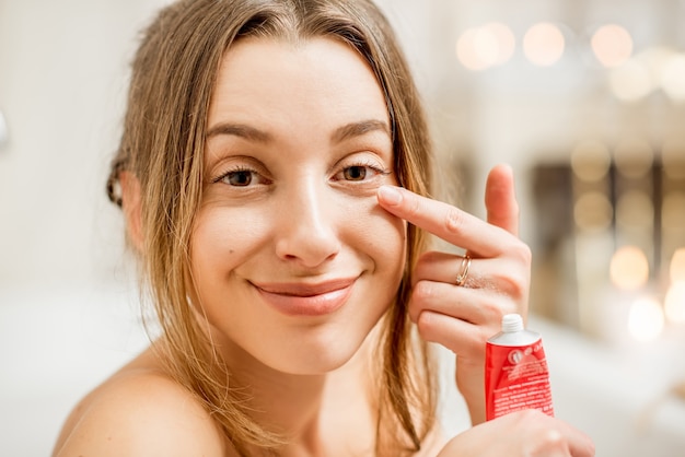 Jeune et jolie femme appliquant une crème faciale sous les yeux dans la salle de bain