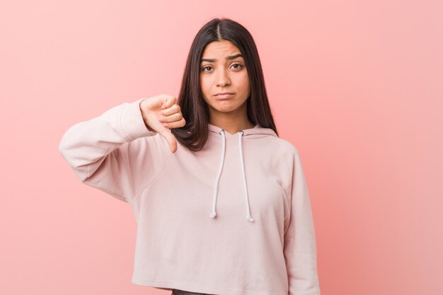 Jeune jolie femme arabe vêtue d'un look sport décontracté, montrant un geste d'aversion, les pouces vers le bas. Notion de désaccord.