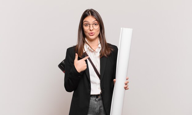 Jeune jolie femme architecte semblant choquée et surprise avec la bouche grande ouverte, pointant vers soi