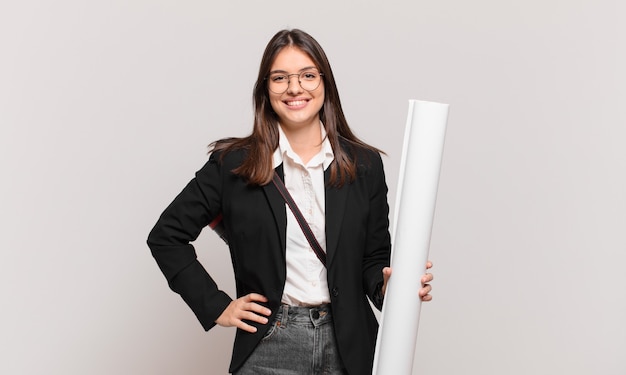 Jeune jolie femme architecte souriant joyeusement avec une main sur la hanche et une attitude confiante, positive, fière et amicale